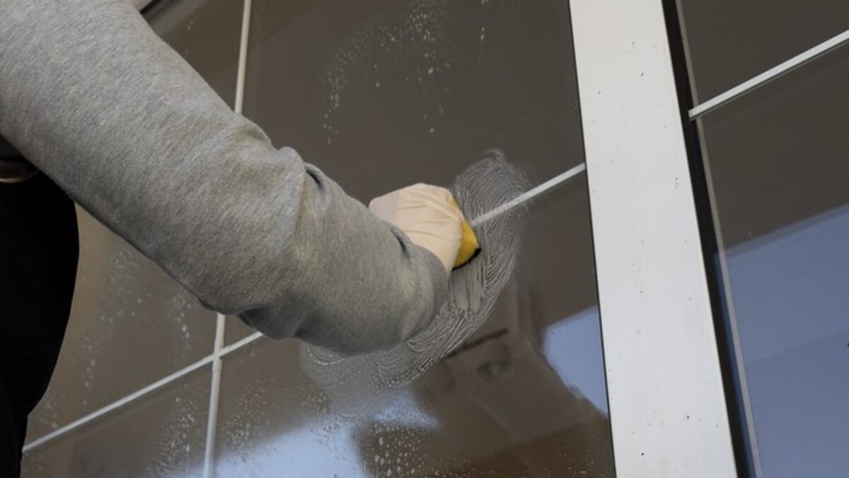 A gloved hand working a soapy cleaning head down the wet glass of a house window, cleaning water running down the pane
