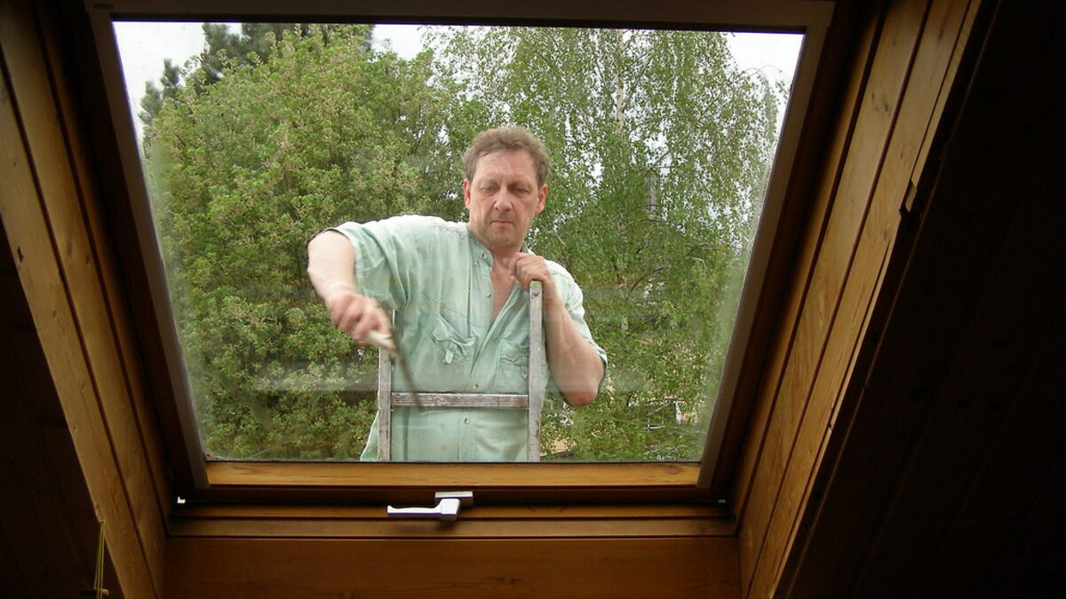 Seen from a timber-beamed room, a window cleaner in a green work shirt stands on a ladder outside and draws a squeegee down the house window